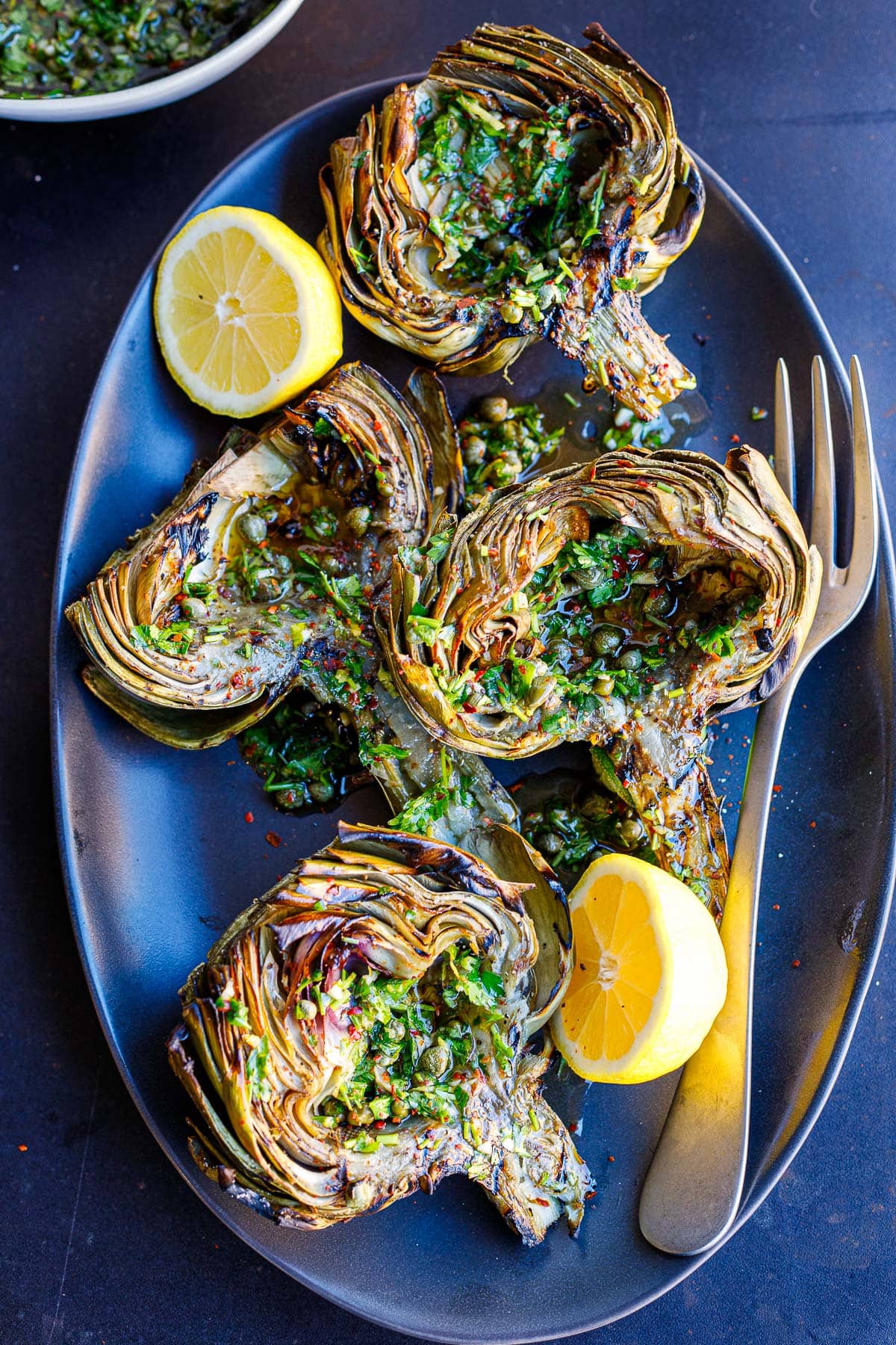 serving plate with grilled artichokes, served with italian salsa verde and lemon halves.