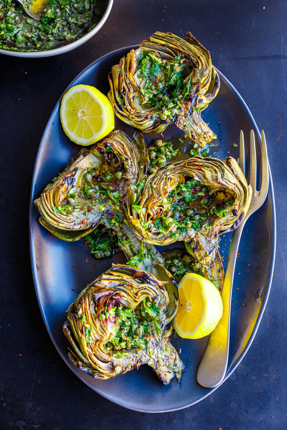 serving plate with grilled artichokes, drizzled with Italian salsa verde, served with lemons.