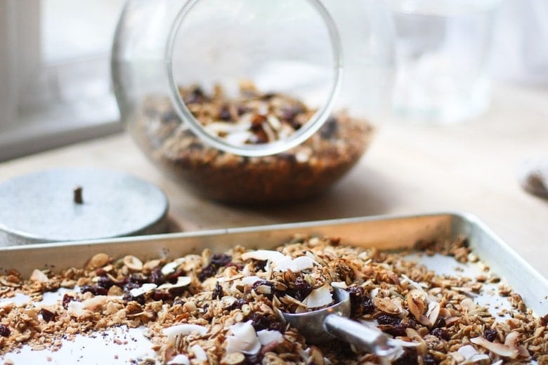 baked homemade granola on baking sheet with dried fruit and coconut flakes mixed in- metal scoop to pour into glass jar.