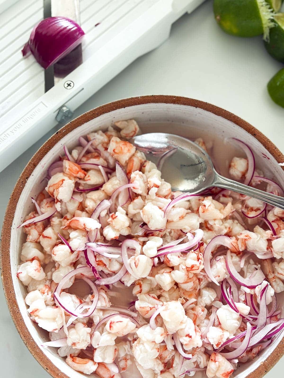 shrimp pieces and sliced red onion in a dish with lime juice and a spoon to make ceviche.