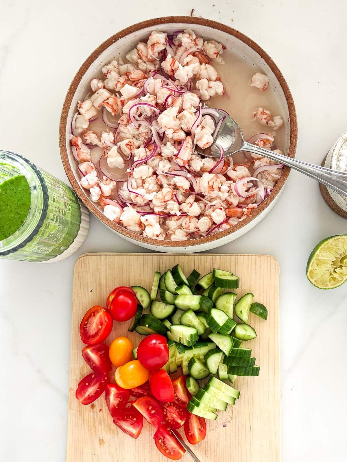 components to make shrimp ceviche verde - jar of verde sauce, bowl of shrimp and red onion in lime juice, wood cutting board with chopped tomato and cucumber.