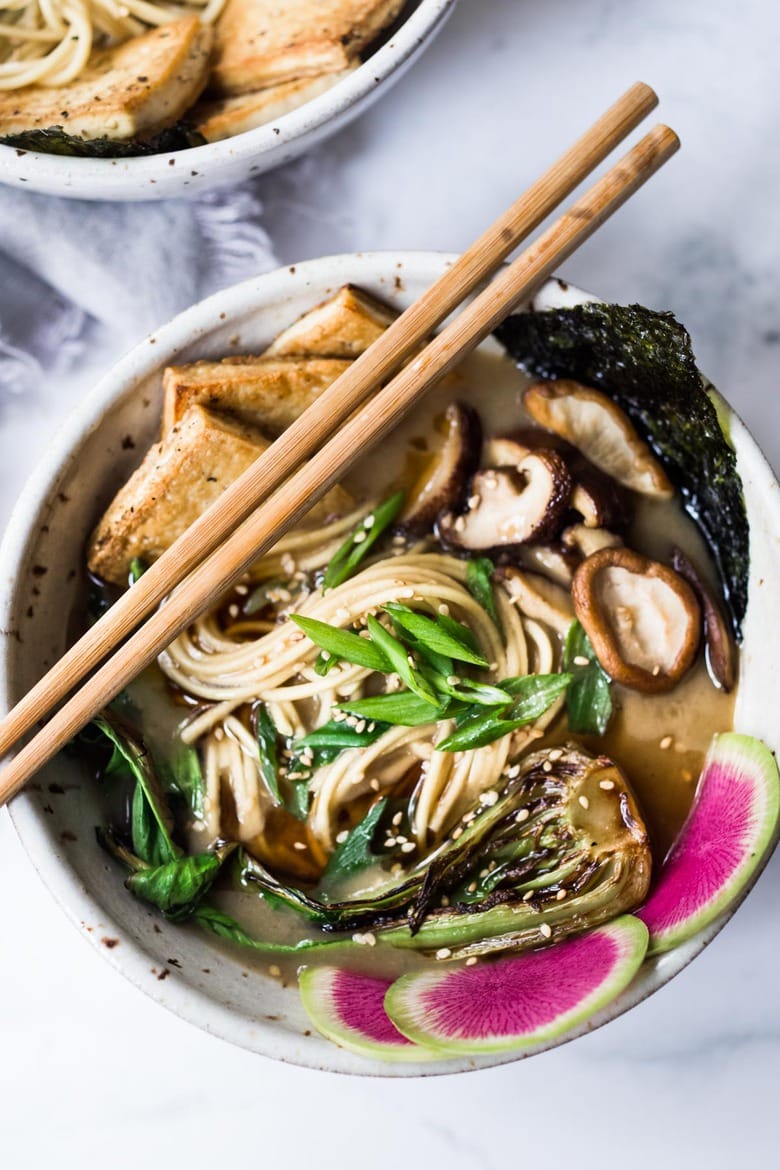 Vegan Ramen bowl of vegan ramen with watermelon radish, bok choy, shiitake mushrooms, crispy tofu, kombu, and scallions with chopsticks on top, garnished with toasted sesame seeds.