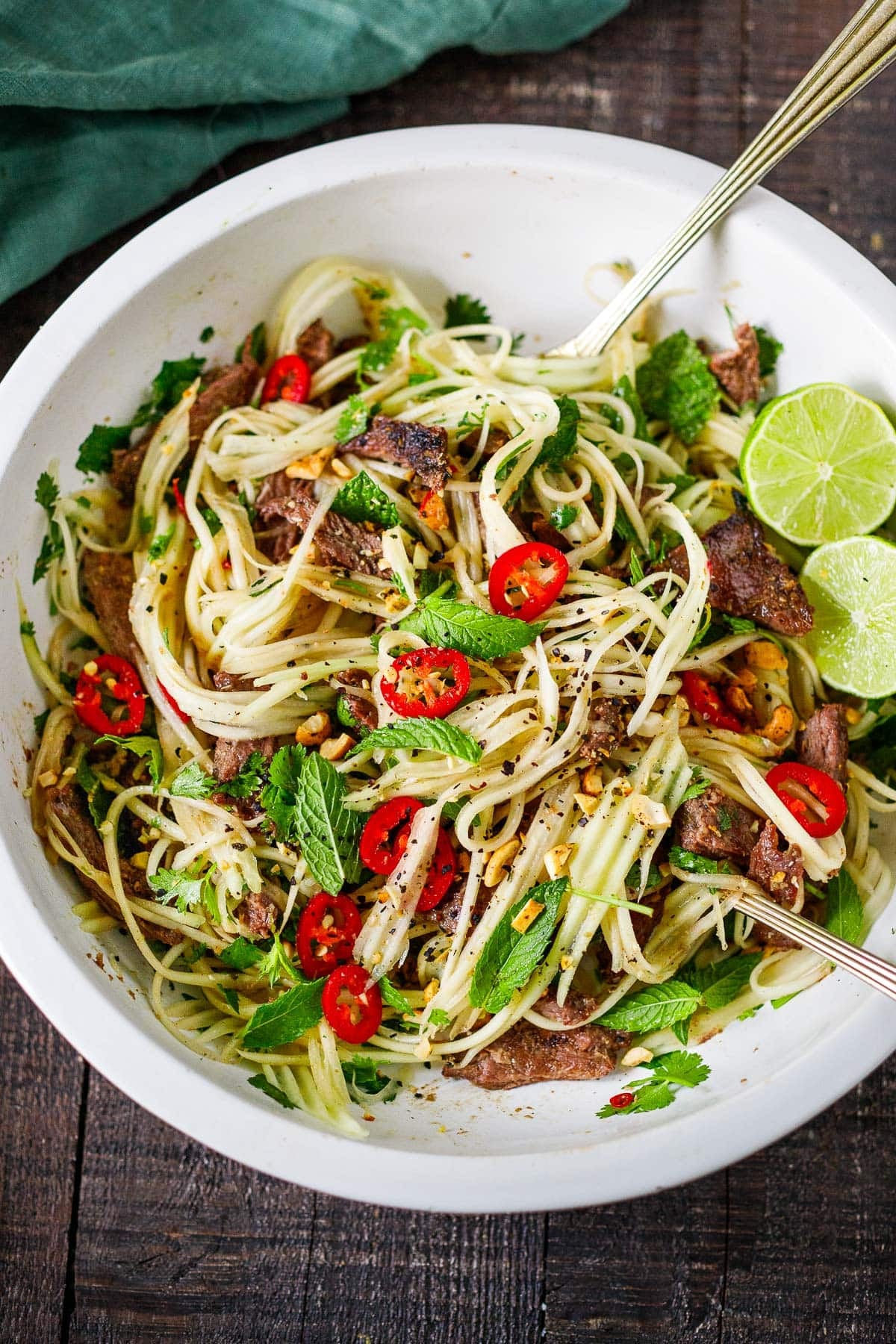green papaya salad with grilled beef, herbs, red chilies, and lime halves in a bowl with serving utensils.