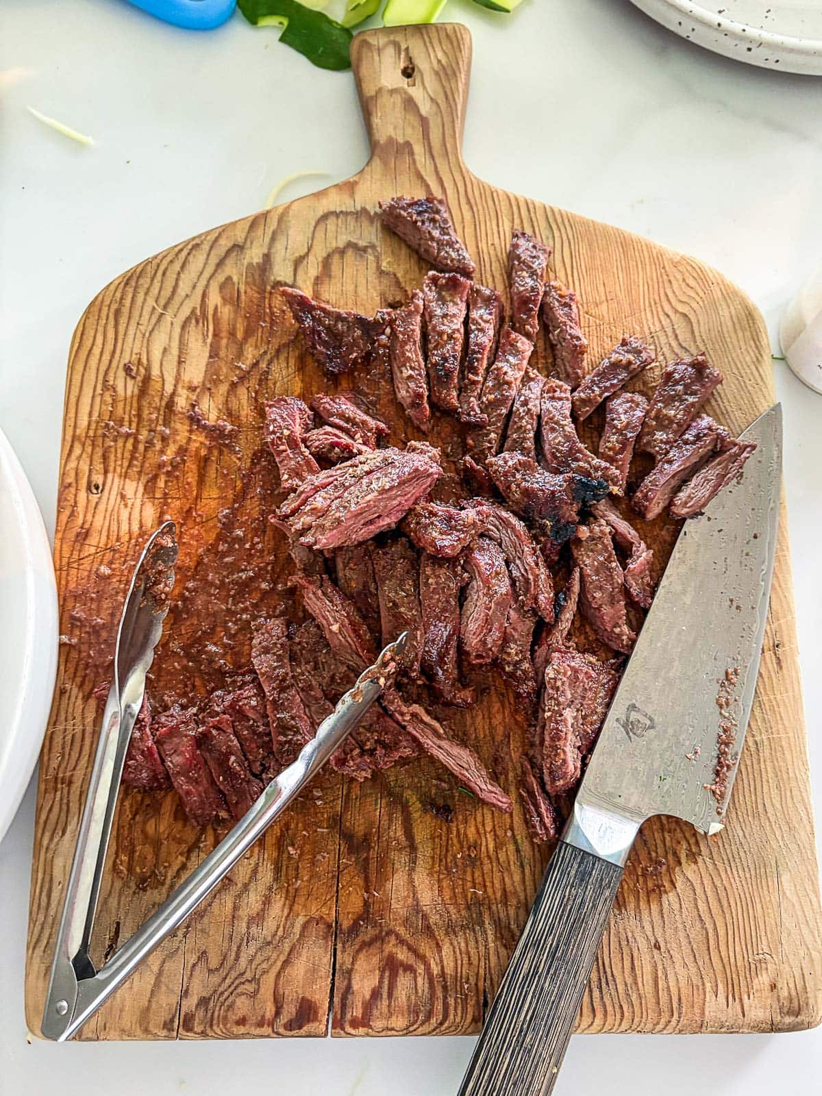 sliced grilled beef on a wood board with a sharp knife and tongs.