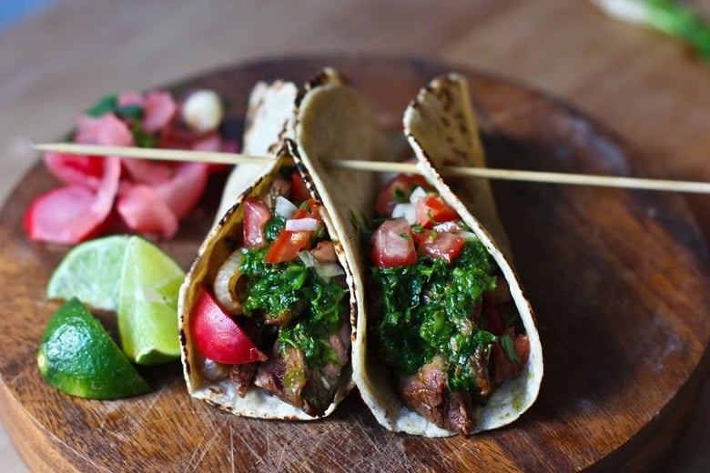 Grilled Steak Tacos with Cilantro Chimichurri Sauce and Pico De Gallo on a cutting board with limes and pickled veggies.