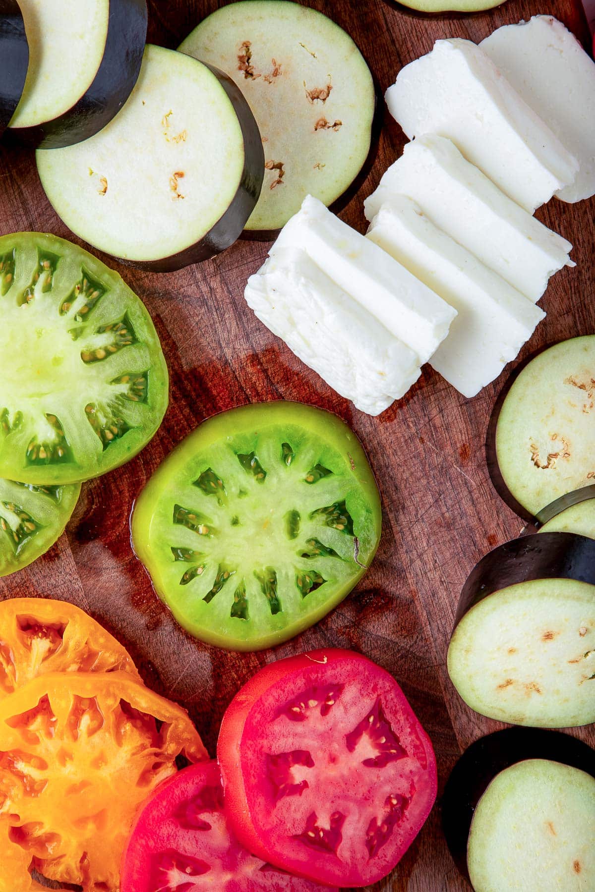 wood cutting board with ingredients for grilled halloumi cheese and eggplant - sliced eggplant, sliced heirloom tomatoes, and sliced halloumi cheese.