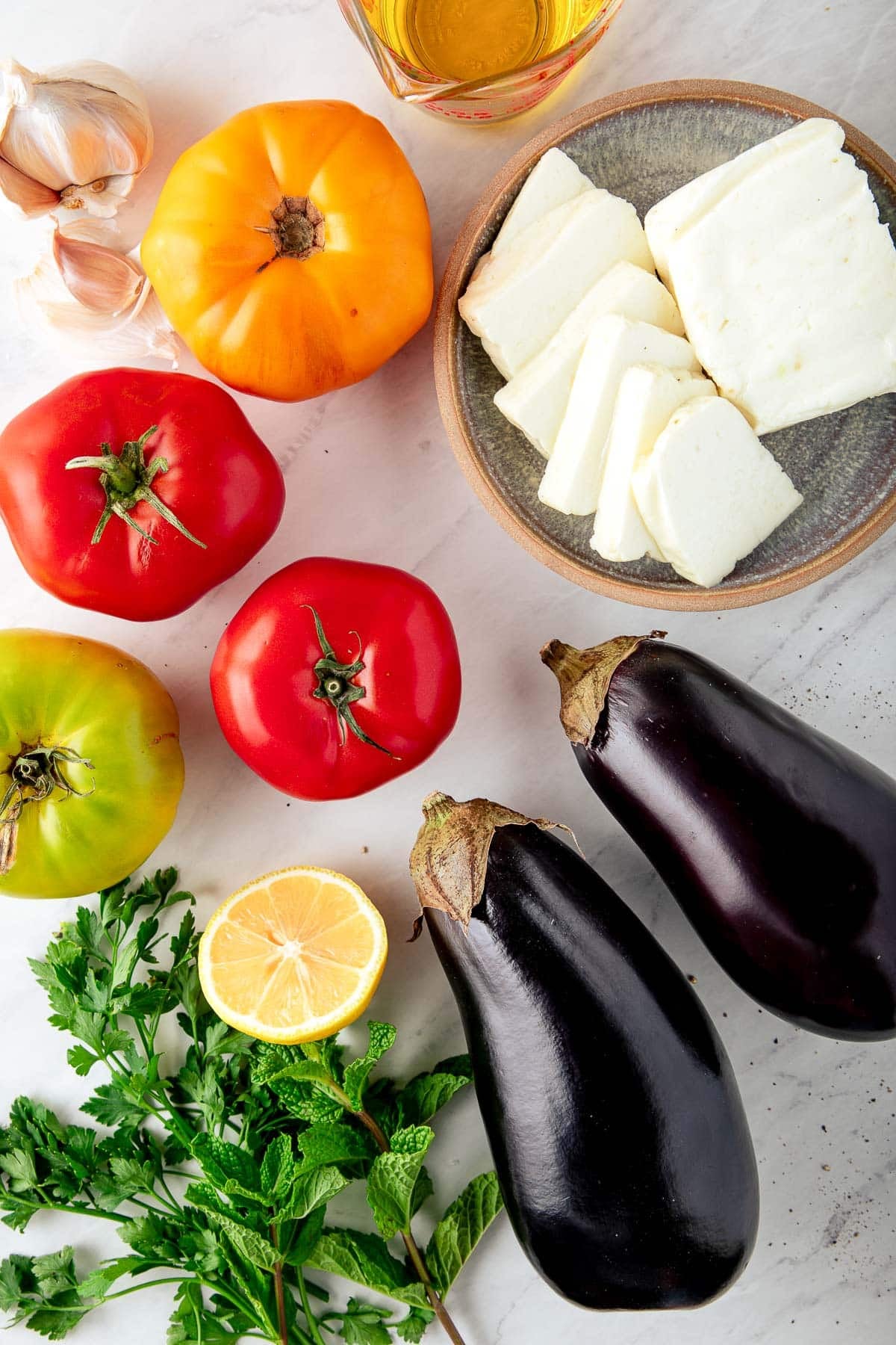 ingredients for grilled halloumi cheese and eggplant - heirloom tomatoes, sliced halloumi cheese, two eggplants, lemon half, and fresh mint and parsley.