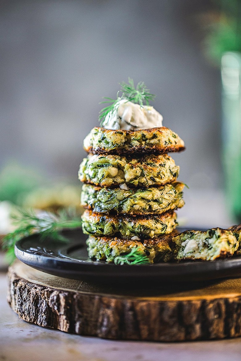 zucchini fritters stacked on plate topped with tzatziki sauce and fresh dill.