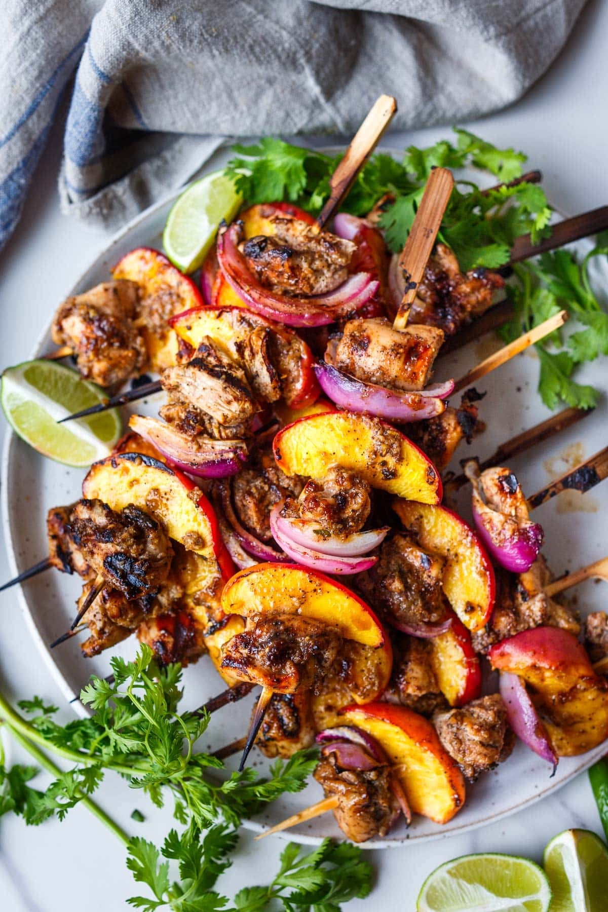 grilled jerk chicken, peach, and red onion skewers stacked on a plate with lime wedge and herbs for a healthy dinner idea for summer.