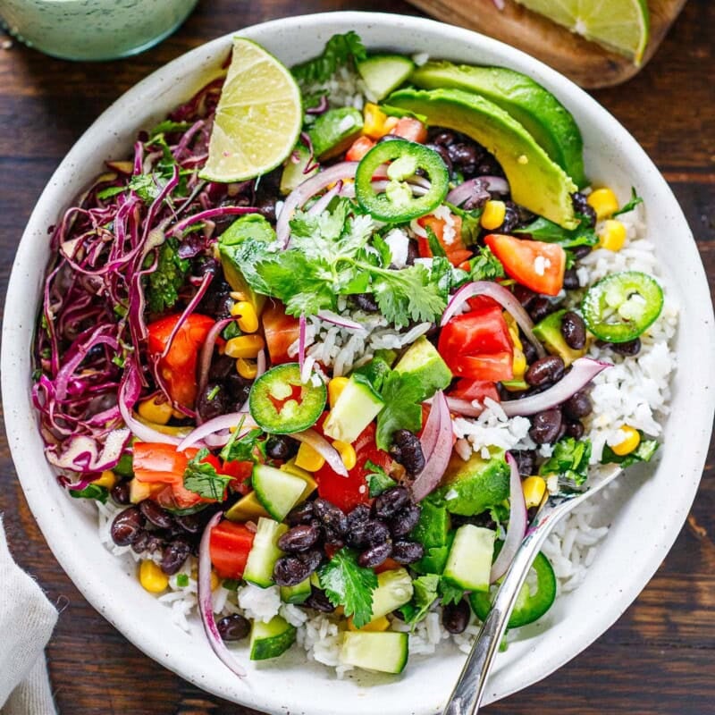 Our favorite burrito bowl recipe is made with seasoned black beans and rice topped with fresh tomatoes, avocado, corn, shredded cabbage, and a vegan cilantro lime crema.