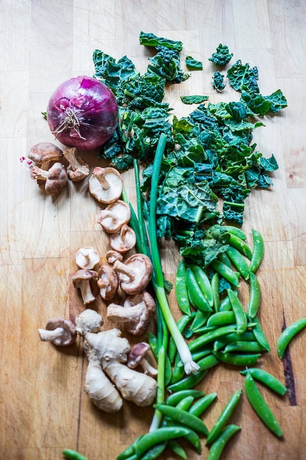 ingredients for Asian noodle soup laid out on wood countertop - kale, red onion, shiitake mushrooms, scallions, snow peas, and fresh ginger.