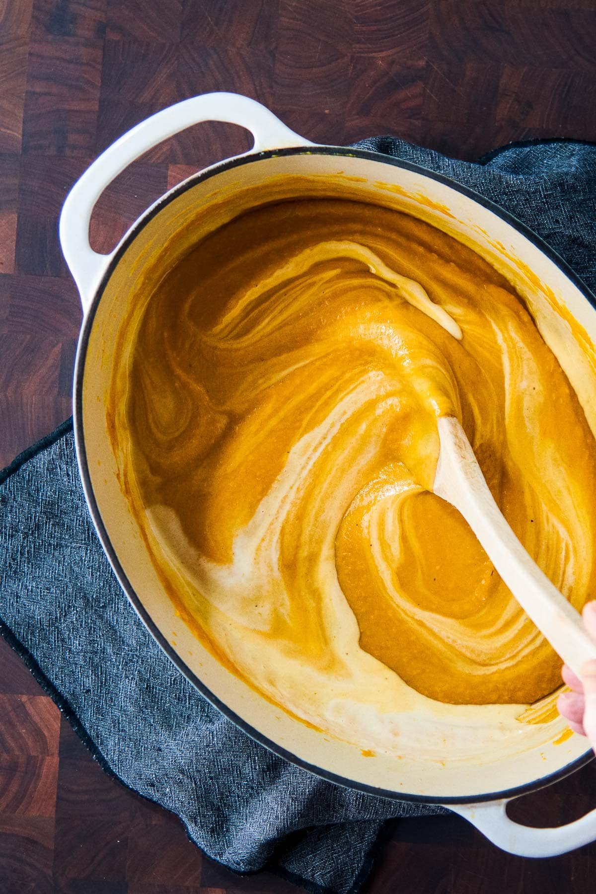 large dutch oven with creamy sweet potato soup with wood spoon swirling in coconut milk. 