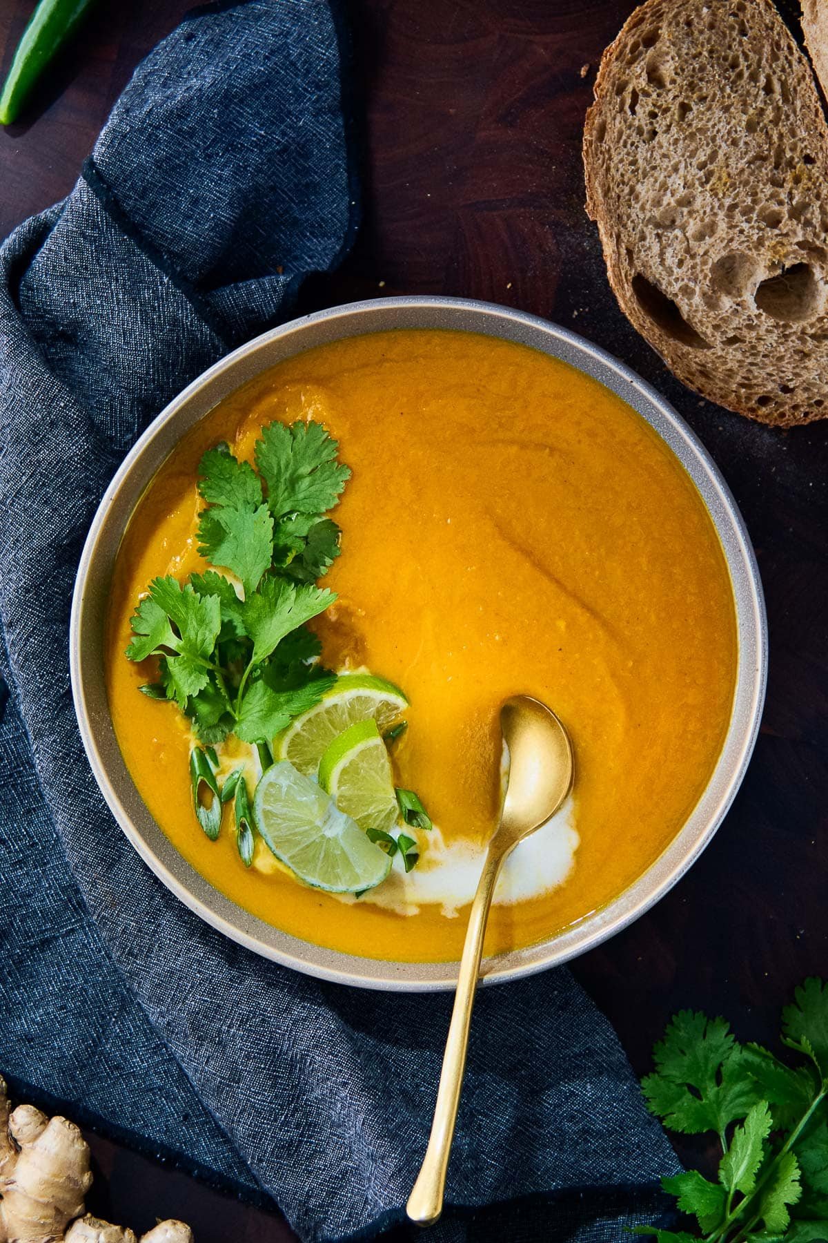 bowl of sweet potato soup with gold spoon and a swirl of coconut milk, garnished with cilantro, green onions, and lime slices. 