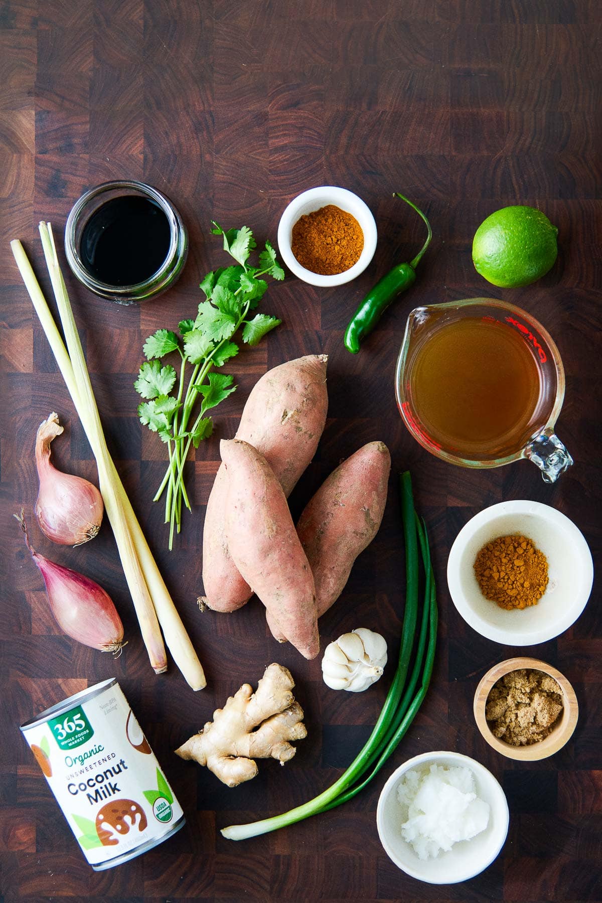 ingredients for sweet potato soup laid out on wood butcher block - soy sauce, cilantro, curry powder, serrano chili, lime, broth, sweet potatoes, turmeric, lemongrass, shallot, garlic, ginger, brown sugar, coconut milk, coconut oil, scallions. 