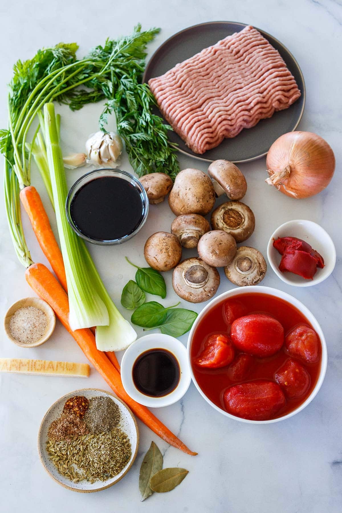 ingredients for turkey bolognese - ground turkey on a plate, onion, mushrooms, garlic bulb, celery stalks, carrots with tops, fresh basil leaves, bowls of red wine, tomato paste, soy sauce, whole canned tomatoes, salt, spices, and Parmesan rind.