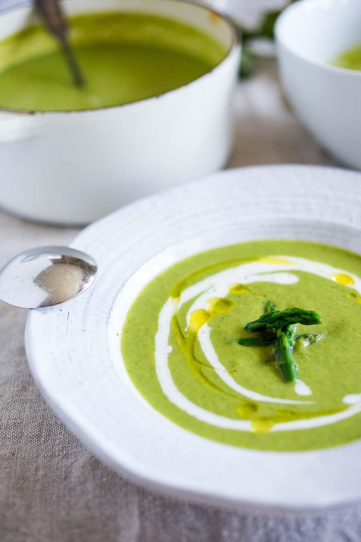 bowl of creamy asparagus soup with swirl of sour cream and oil with a few tender asparagus tips in center.