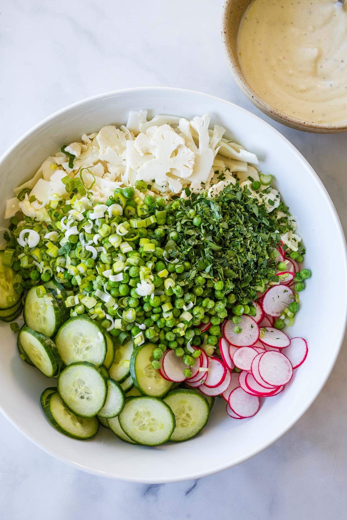all components for raw cauliflower salad in a large salad bowl - sliced cauliflower, radish, and cucumber, peas, herbs, scallions.