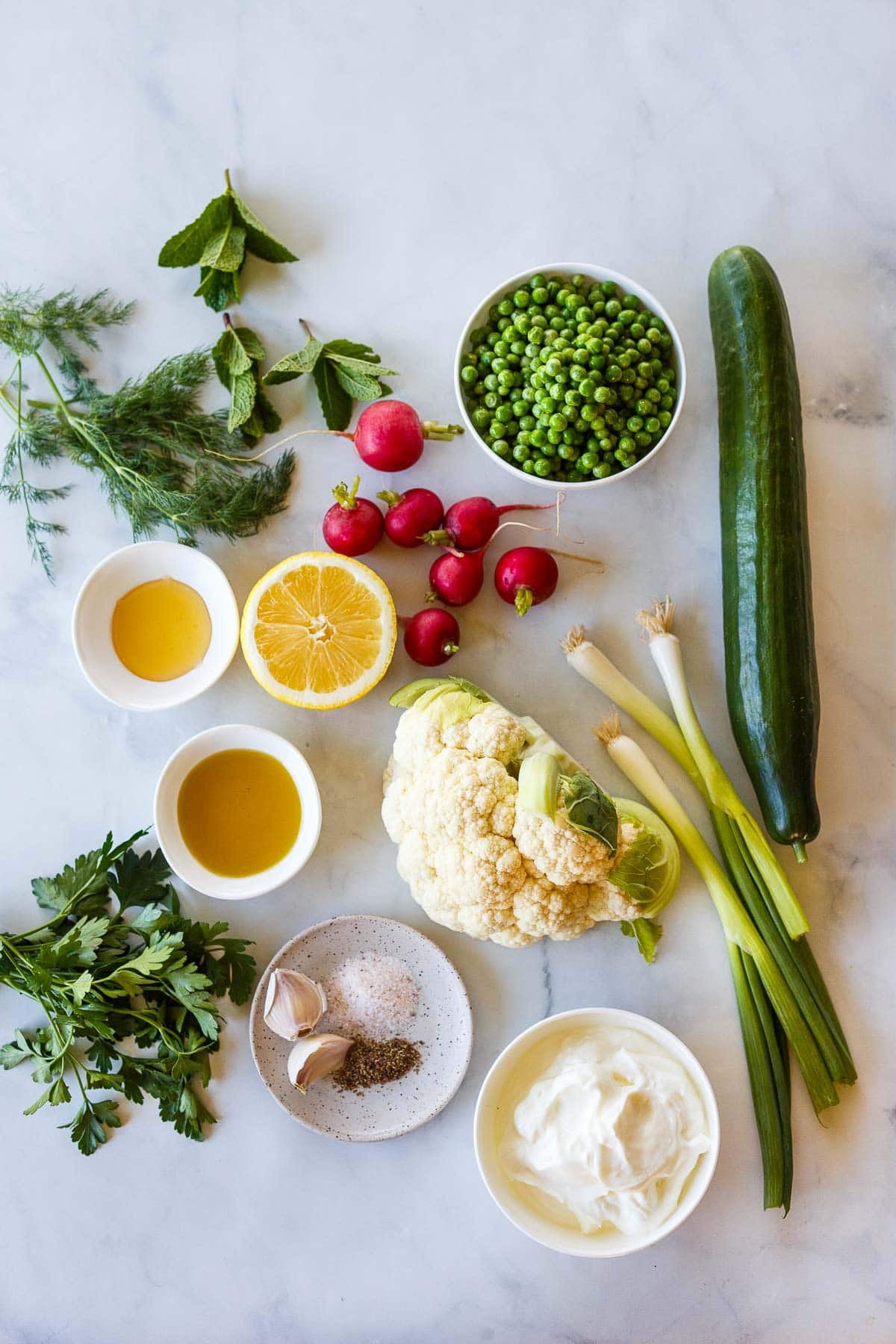 ingredients for cauliflower salad - dill, mint, peas, radishes, cucumber, lemon half, honey, oil, cauliflower head, green onions, parsley, garlic cloves, salt, pepper, Greek yogurt.