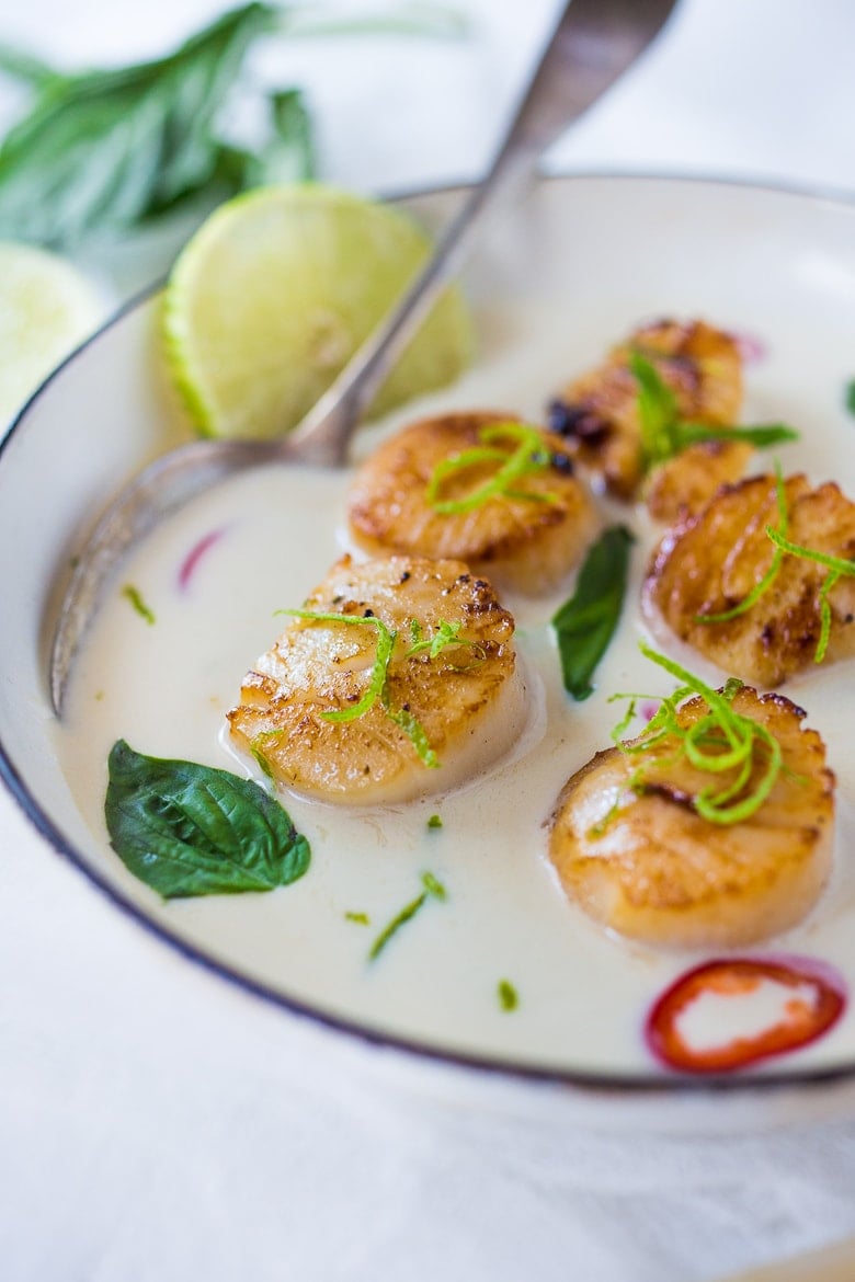 bowl of seared scallops in coconut lemongrass sauce topped with basil leaves, lime zest, and red chilis.