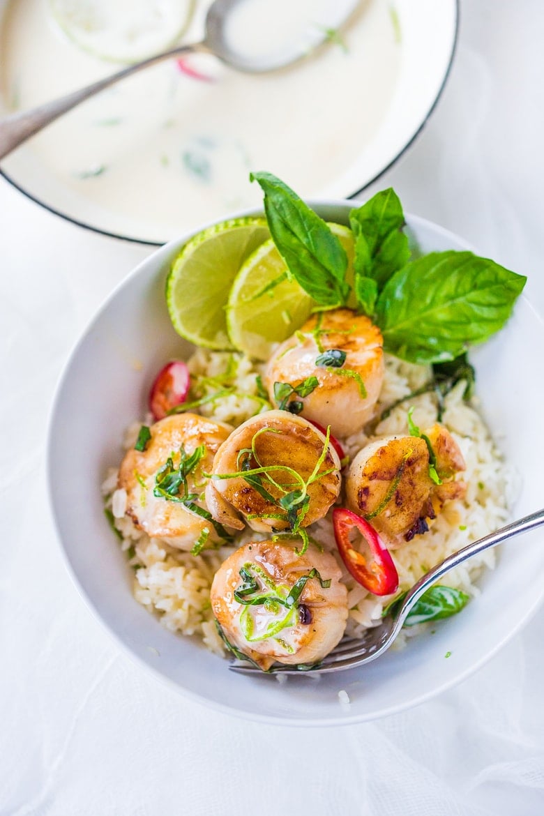 bowl of rice with seared scallops with coconut lemongrass sauce, garnished with lime zest, basil ribbons, and sliced chilis, with fork.