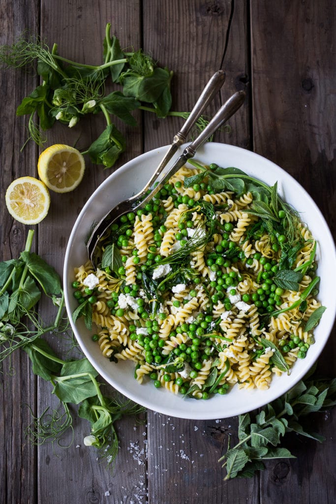 spring pea pasta salad with fresh herbs and goat cheese in white serving bowl with serving spoons.