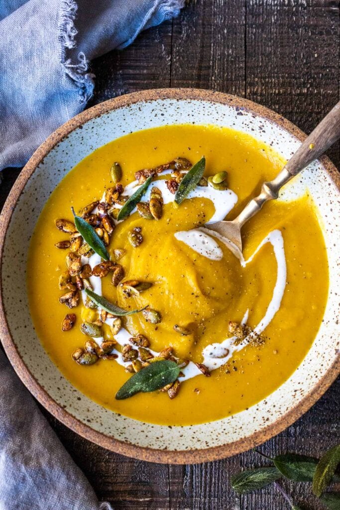 bowl of butternut squash soup with coconut milk drizzle, toasted pepitas, and sage leaves with spoon.