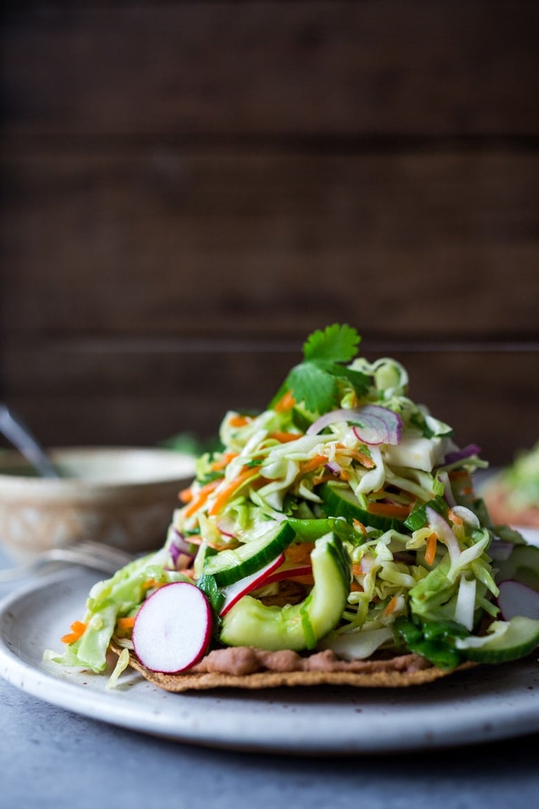 Introducing Tlayudas! Traditional Oaxacan street food, tlayudas are a cross between a tostada and a pizza, made with a crispy tortilla, topped with refried beans, cheese (optional) cabbage slaw, avocado and cilantro. This healthy version is vegan and GF adaptable! | #tlayudas #tostadas #mexicanpizza #vegan #oaxacanfood #mexicanstreetfood #veganmexican #healthymexicanrecipes | www.feastingathome.com