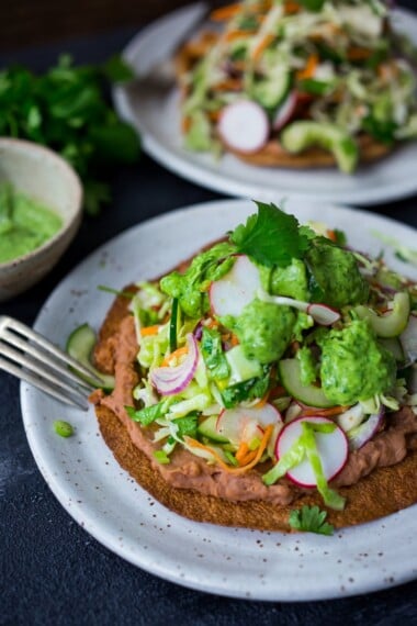 Introducing Tlayudas! Traditional Oaxacan street food, tlayudas are a cross between a tostada and a pizza, made with a crispy tortilla, topped with refried beans, cheese (optional) cabbage slaw, avocado and cilantro. This healthy version is vegan and GF adaptable! | #tlayudas #tostadas #mexicanpizza #vegan #oaxacanfood #mexicanstreetfood #veganmexican #healthymexicanrecipes | www.feastingathome.com