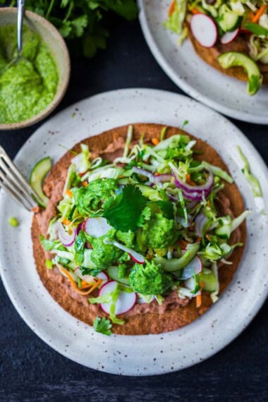 How to make Oaxcan Tlayudas! Oaxacan street food, tlayudas are a cross between a tostada and a pizza, made with a crispy tortilla, topped with refried beans, cheese (optional) cabbage slaw, avocado and cilantro. This healthy version is vegan and GF adaptable!