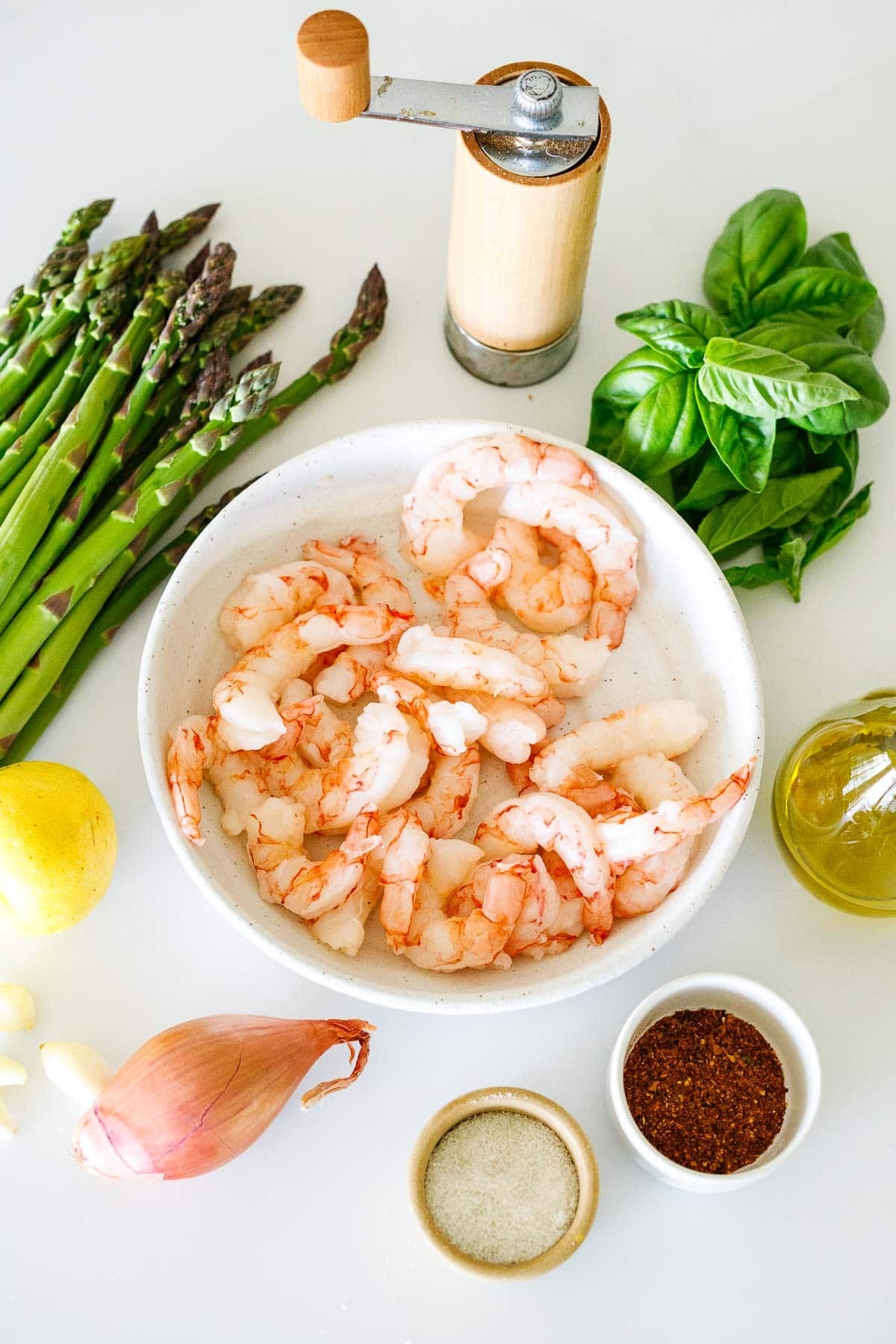 one pan shrimp and asparagus ingredients - pepper shaker, basil leaves, asparagus bunch, bowl of shrimp, oil, chili flakes, salt, shallot, garlic cloves, and lemon.