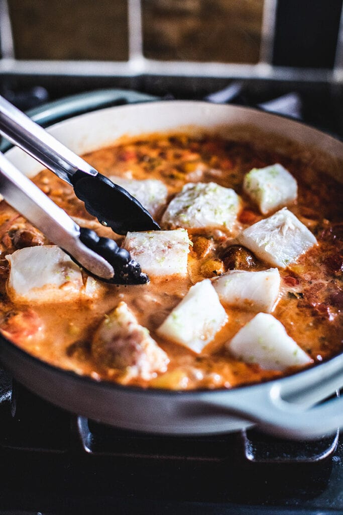 tongs placing fish fillets into pan of Moqueca, a Brazilian fish stew. 