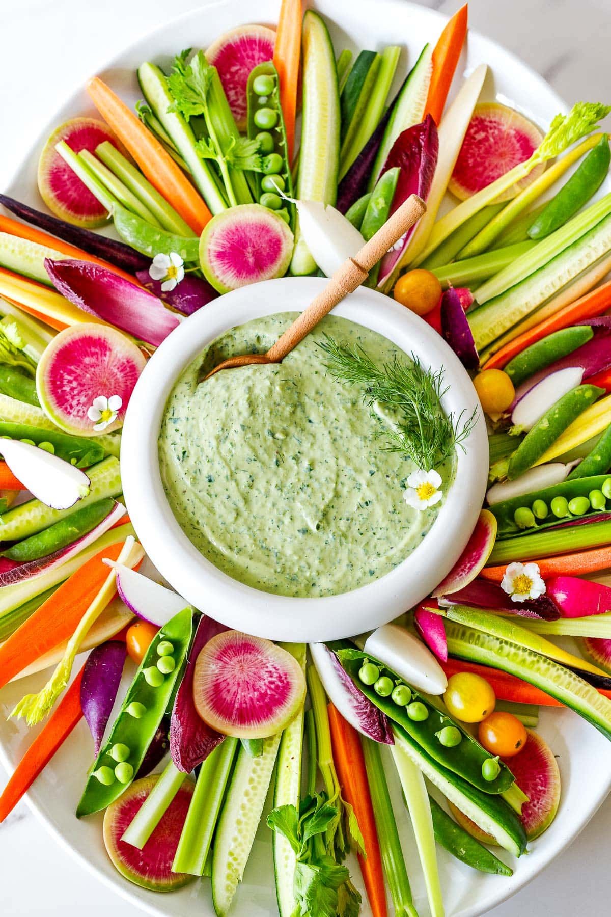 oval crudite platter with colorful vegetables in strips around a white bowl of green goddess dressing topped with dill and an edible flower with a wood spoon.