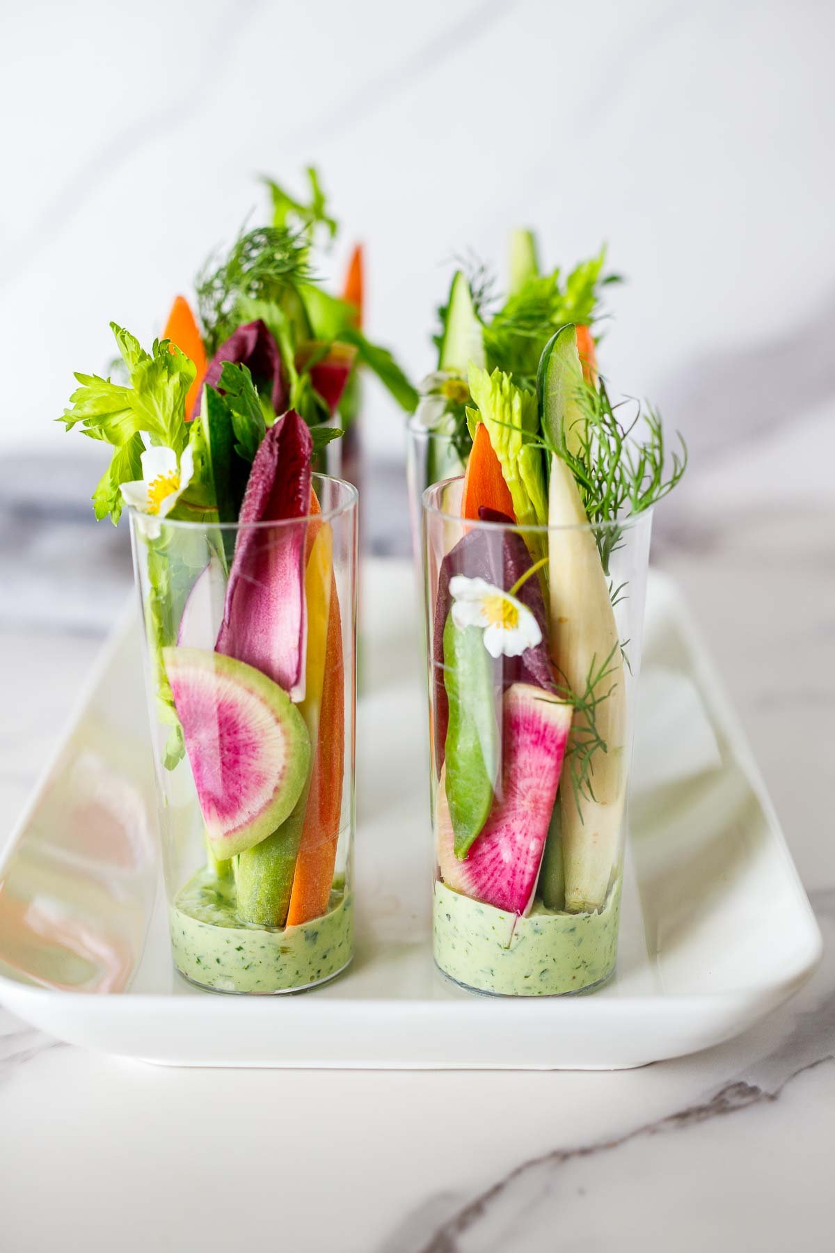 catering shot glasses on a rectangular tray with green goddess dressing in the bottom and thinly sliced veggies placed on top.