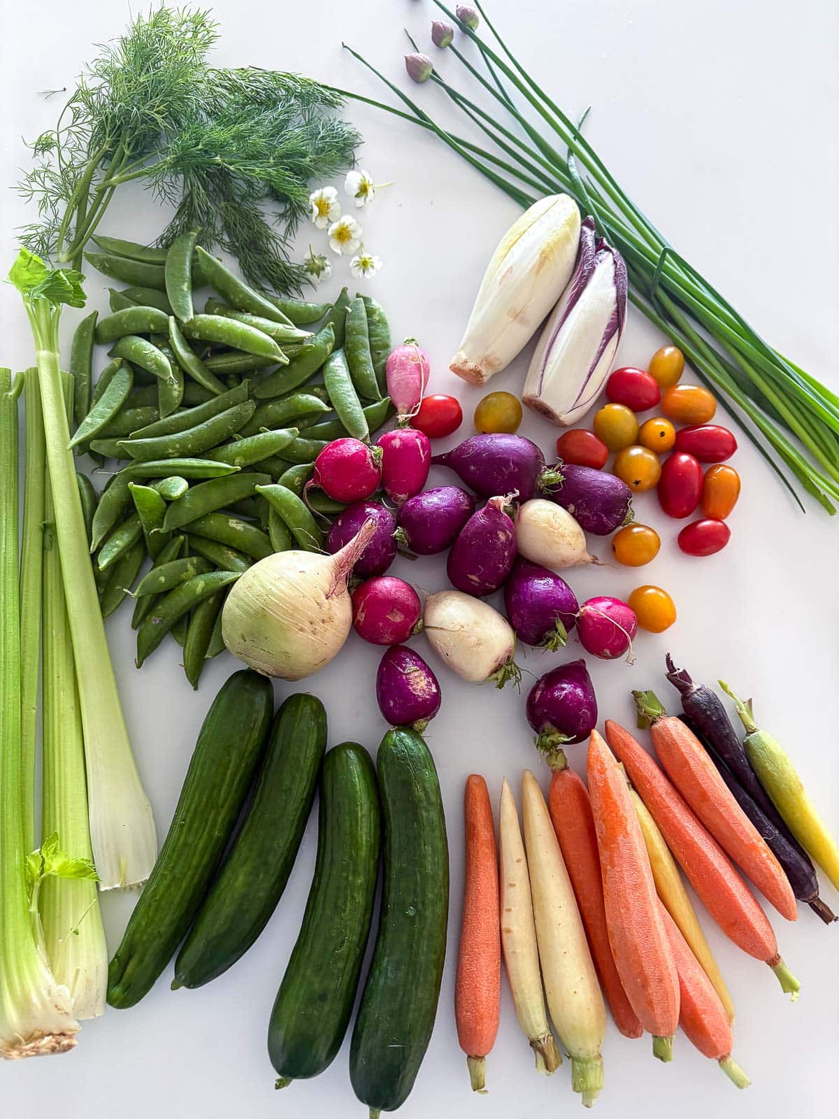 colorful vegetables for crudite arranged on counter - chive blossoms, endive, cherry tomatoes, radishes, sugar snap peas, dill, edible flowers, celery, carrots, cucumber.