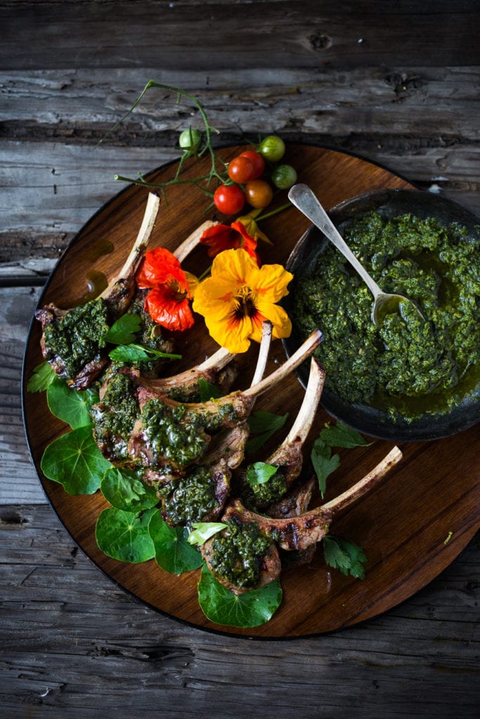 grilled lamb chops with Italian salsa verde on a round wood platter with flowers, tomatoes, and herbs.