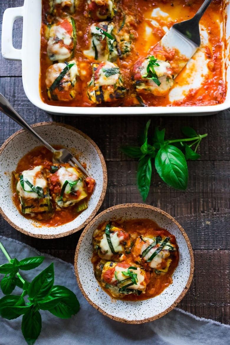 baking dish with metal spatula serving zucchini lasagna roll ups and two bowls with roll ups topped with melty cheese, marinara, and basil.