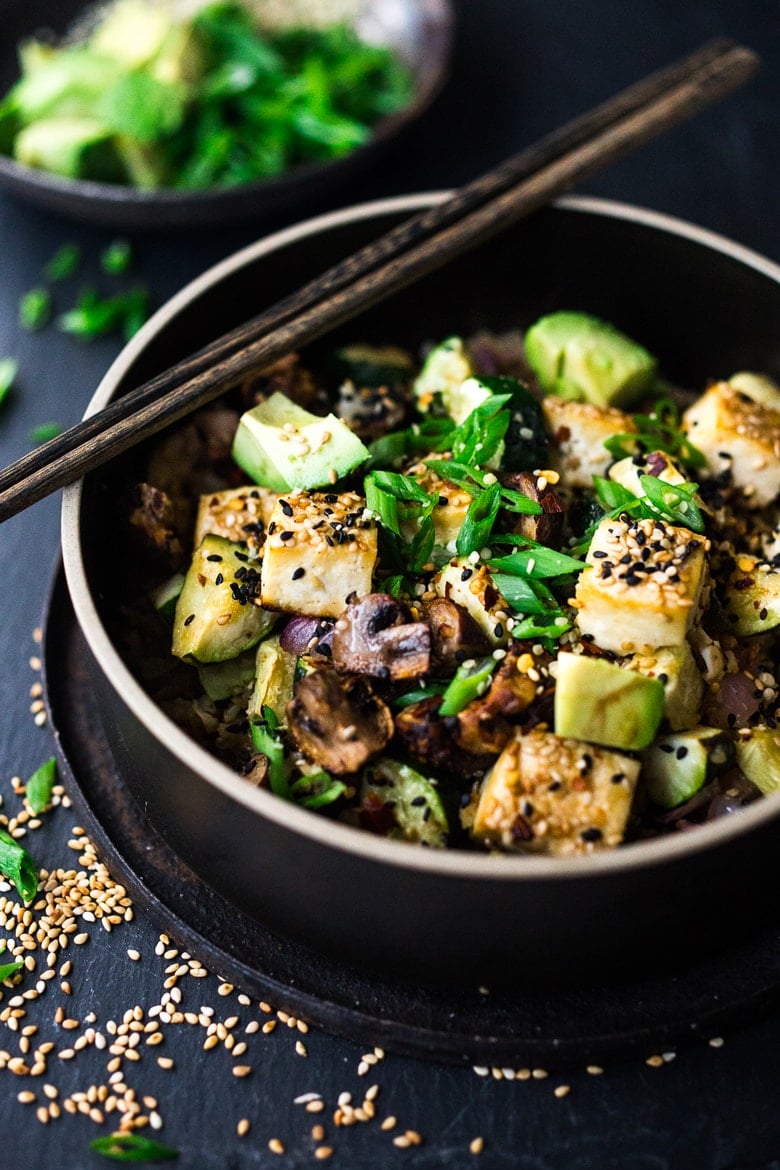 cauliflower rice bowl with roasted mushrooms and cubed baked tofu and cubed avocado garnished with sesame seeds and green onions with chopsticks on the bowl.