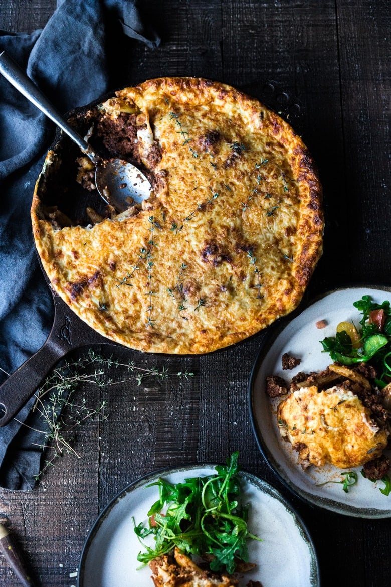 cast iron skillet with baked Greek lamb pasta, beside two plates with a serving of pastitsio and side salad.