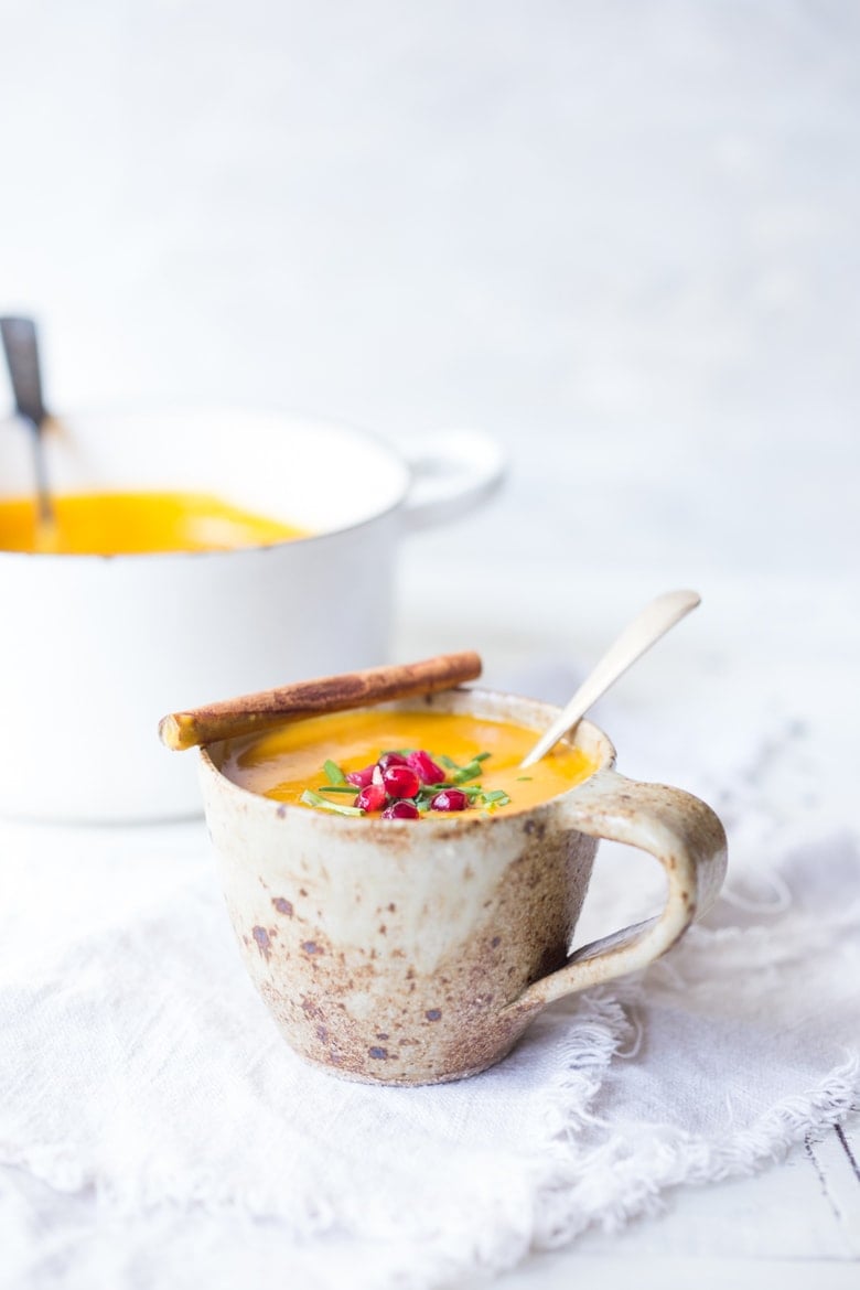 Moroccan carrot soup in mug with spoon, served with pomegranate seeds and chives, garnished with cinnamon stick.