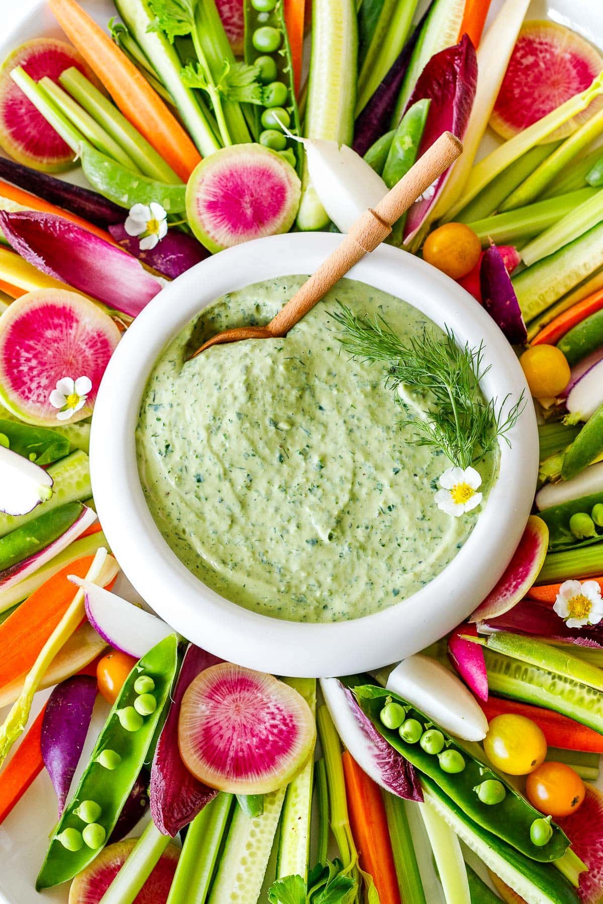 crudite platter of colorful vegetables arranged neatly around a bowl of green goddess dip with wood spoon, garnished with dill and white flower.