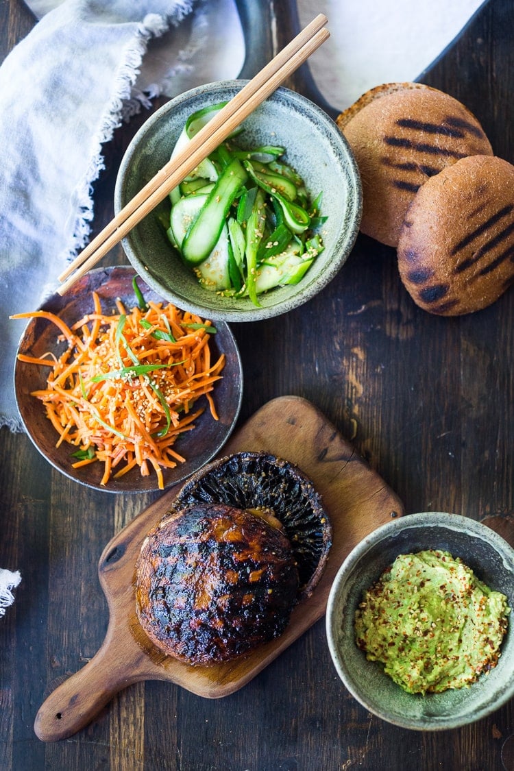 marinated and grilled portobello mushrooms beside bowls with carrot slaw, cucumber ribbon salad, and Asian guacamole.