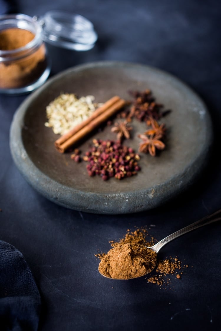spoonful of homemade Chinese Five Spice in front of dish with whole spices used to make it- szechuan peppercorns, star anise, cloves, cinnamon stick, fennel seeds.