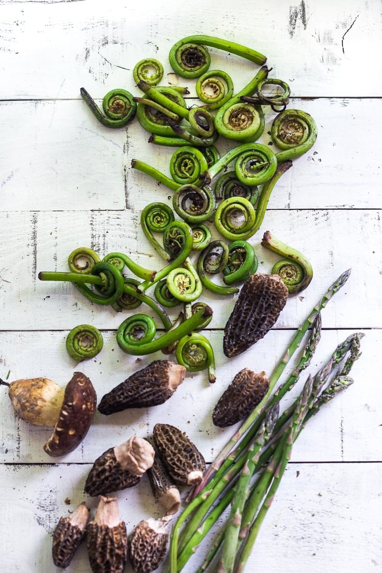 spring veggies laid out on wood table - fiddlehead ferns, mushrooms, asparagus.