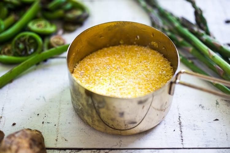 measuring scoop with ground dry polenta with spring veggies in background.