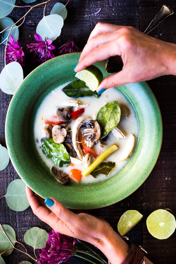 hands squeezing lime quarter into bowl of Tom Kha Gai (Thai Coconut Chicken Soup), full of chicken, mushrooms, chilies, lemongrass, kaffir lime leaves in coconut broth.