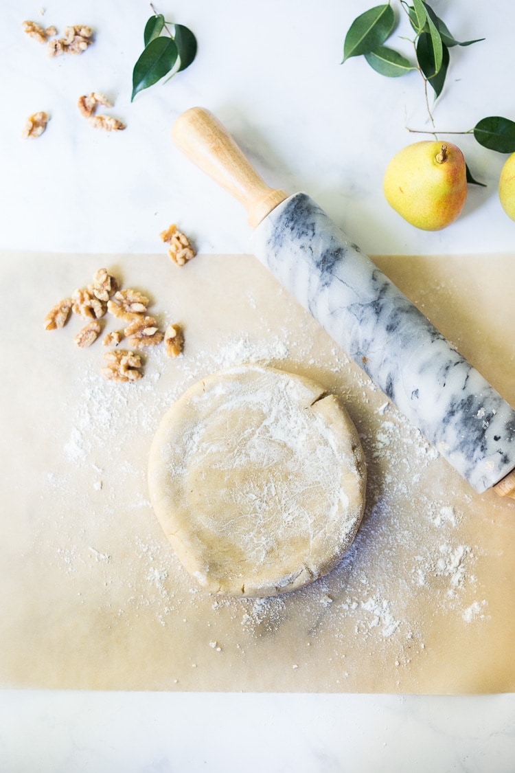 galette dough in disk shape on floured parchment with marble rolling pin and pile of walnuts.