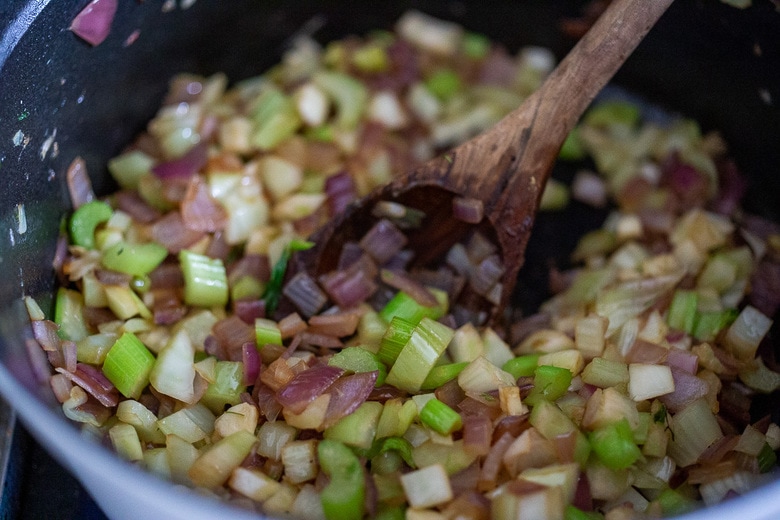 wood spoon stirring sautéed onions, celery, and fennel in pot.