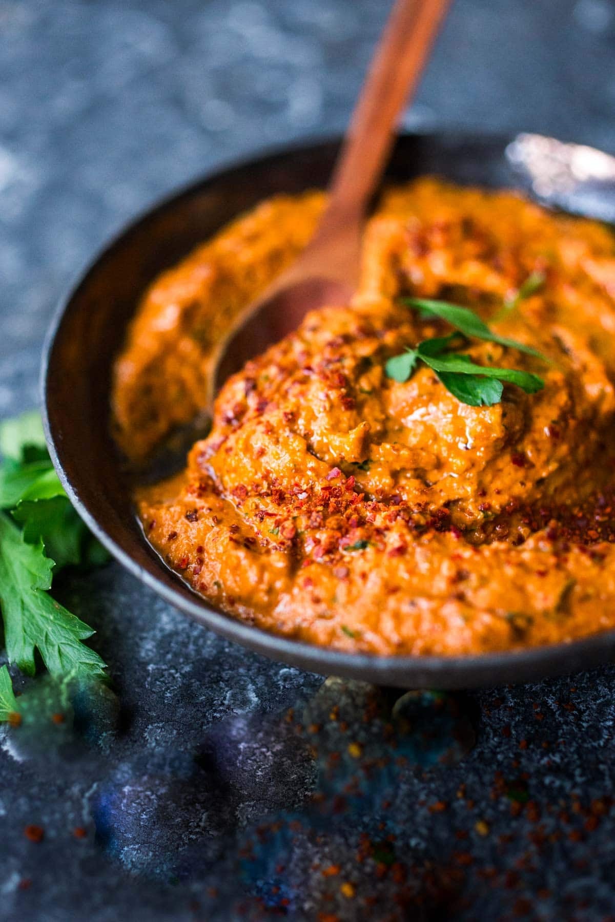 romesco sauce in a bowl with a fresh herb garnish and a spoon, on a wood board with almonds. 