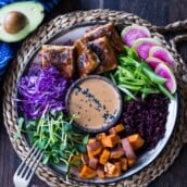 our best buddha bowl recipes - bali bowl with peanut tofu, sliced radish, green onions, black rice, sliced avocado, roasted sweet potato, microgreens, and shredded cabbage surrounding a small bowl of peanut sauce with sesame seeds.