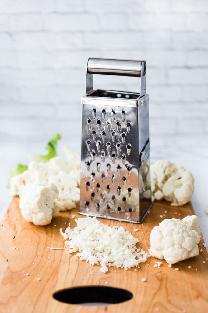 box grater and pieces of cauliflower and grated cauliflower on wood board.